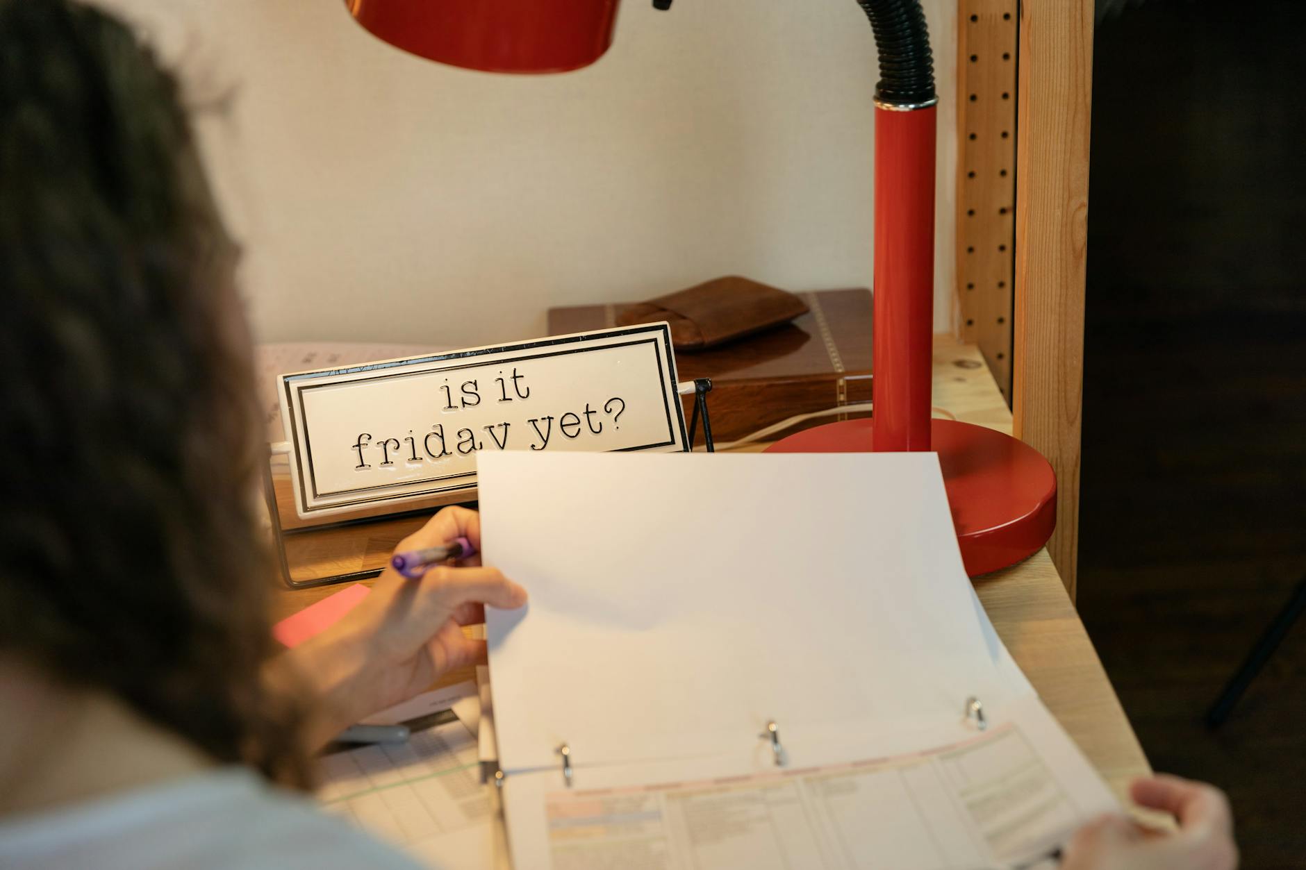 Cozy home office setup with red desk lamp, sign 'Is it Friday yet?', and person looking at documents.