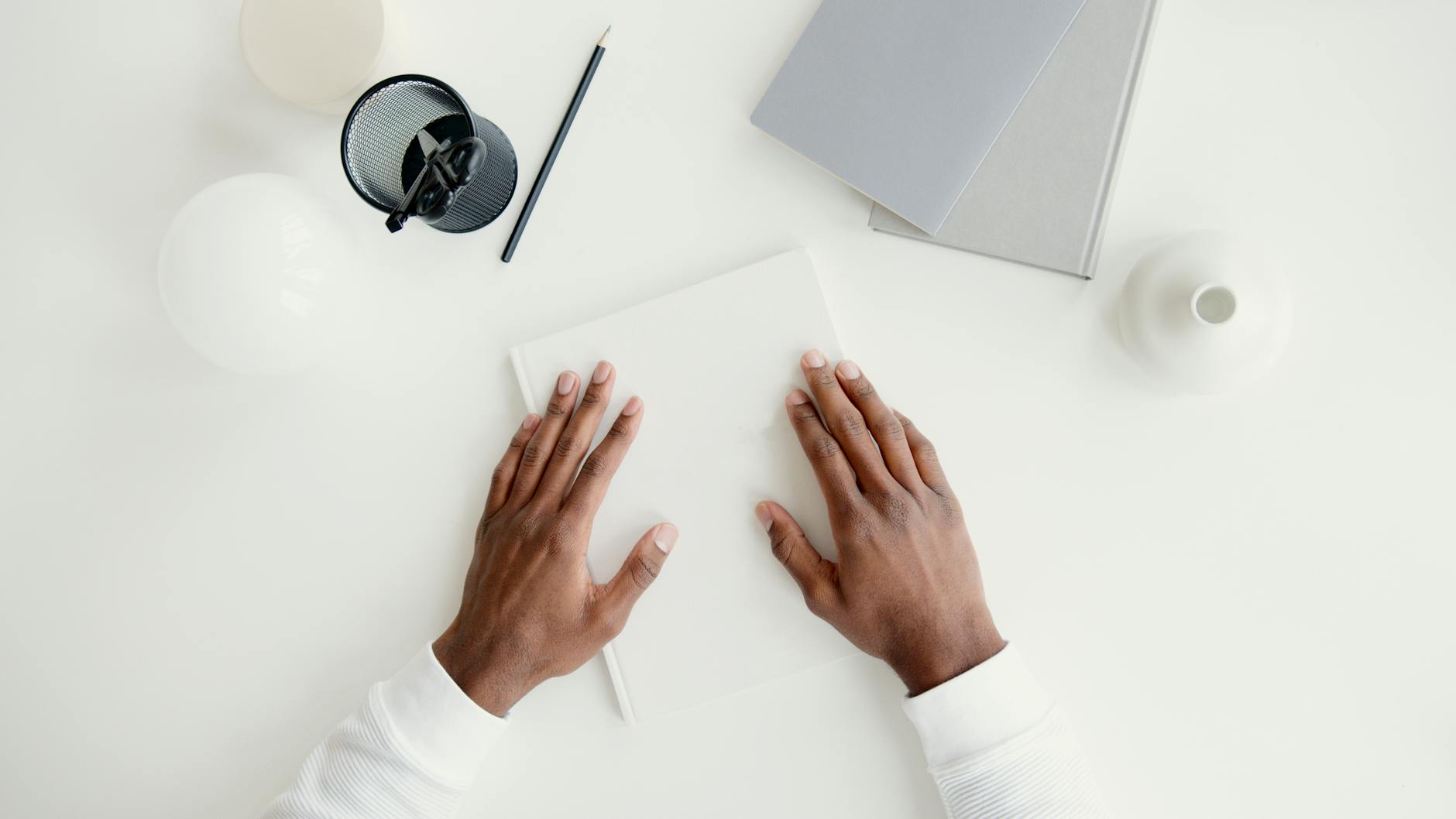 A pair of hands on a minimalist white desk with stationery and books, ideal for business or office concepts.